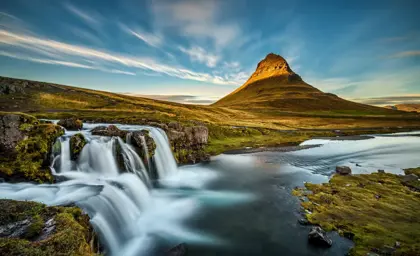 A breathtaking view of Kirkjufellsfoss waterfall, seen at a Snæfellsnes peninsula private tour, cascading in the foreground with the iconic Kirkjufell mountain illuminated by golden sunset light on Snæfellsnes peninsula in Iceland.