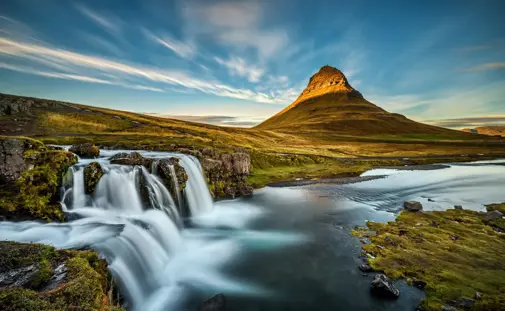 A breathtaking view of Kirkjufellsfoss waterfall, seen at a Snæfellsnes peninsula private tour, cascading in the foreground with the iconic Kirkjufell mountain illuminated by golden sunset light on Snæfellsnes peninsula in Iceland.