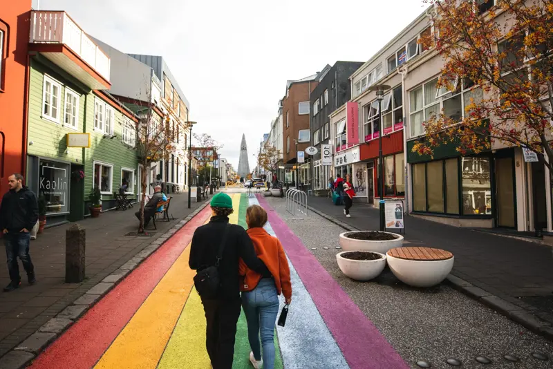 A couple walking along the colorful rainbow-painted street leading to Hallgrímskirkja in Reykjavík, Iceland, surrounded by vibrant buildings and autumn trees. Part of a Reykjavík city tour experience.