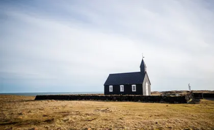 Búðakirkja, the iconic black church surrounded by golden fields and the vast Icelandic coastline, under a bright and serene sky.