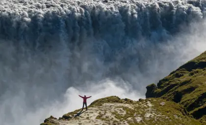 Solo woman in red jacket with arms raised in front of massive cascading Dettifoss waterfall in northeast Iceland