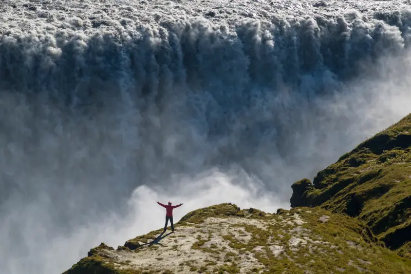Solo woman in red jacket with arms raised in front of massive cascading Dettifoss waterfall in northeast Iceland