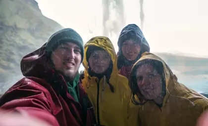 Group of friends in colorful raincoats taking a selfie behind Seljalandsfoss waterfall, enjoying the misty adventure on a South Coast Iceland tour.