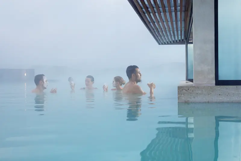 Visitors enjoying the geothermal waters of Blue Lagoon in Iceland with silica face masks, surrounded by steam and minimalistic architecture in a serene spa setting.