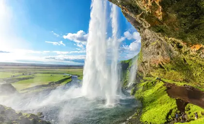 View from behind Seljalandsfoss waterfall, Iceland, showcasing the water stream against a green landscape under a bright blue sky.