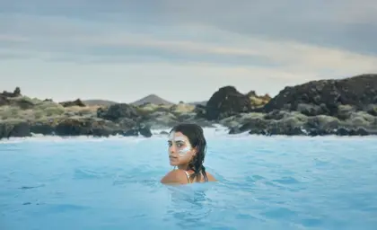 Woman with a white silica face mask relaxing in the Blue Lagoon, a geothermal spa in Iceland, surrounded by volcanic rocks and distant hills under a cloudy sky.