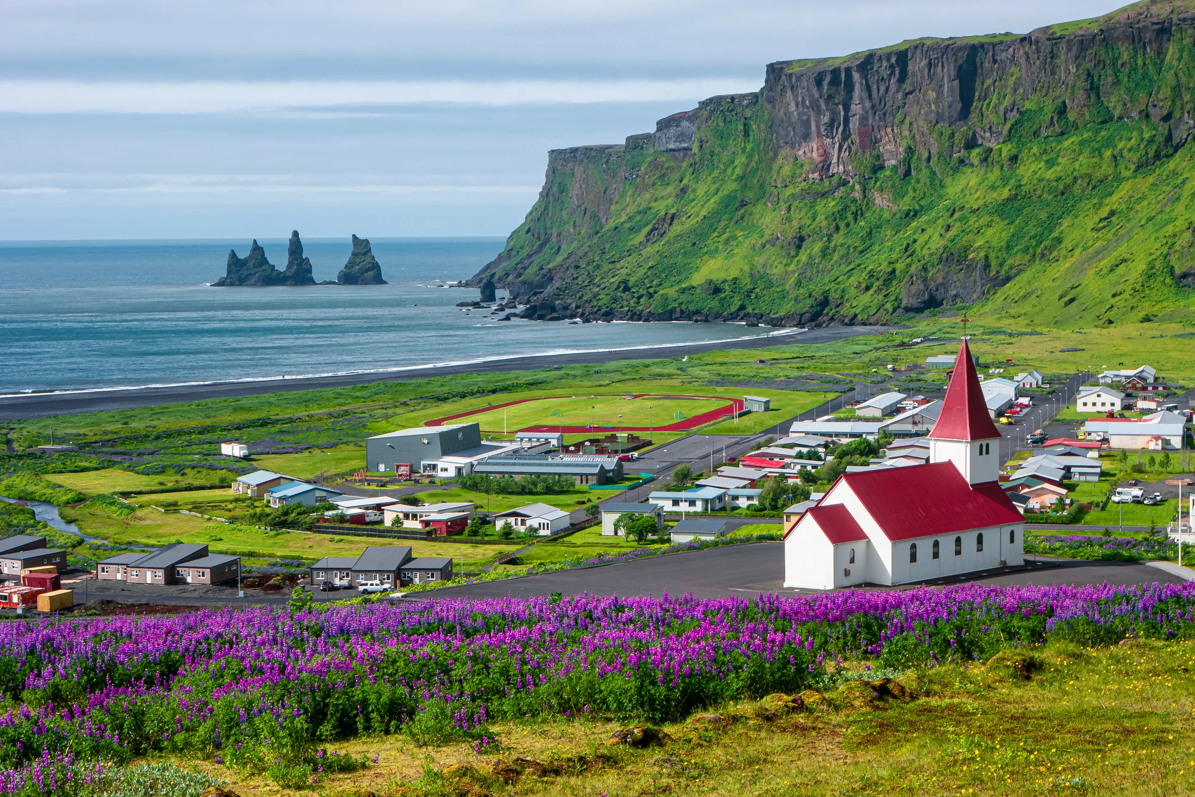 Scenic view of Vík í Mýrdal, showcasing colorful houses, a church with a red roof, and Reynisdrangar sea stacks in the background, highlighting Iceland's natural beauty.