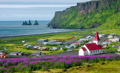 Scenic view of Vík í Mýrdal, showcasing colorful houses, a church with a red roof, and Reynisdrangar sea stacks in the background, highlighting Iceland's natural beauty.
