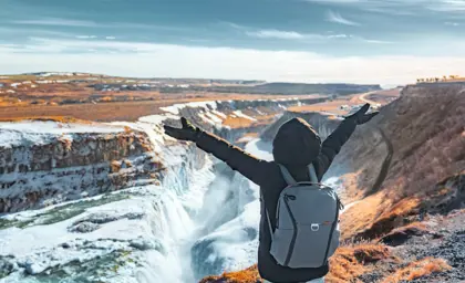 Joyful female tourist raising her arms while admiring the powerful Gullfoss waterfall surrounded by snow in Iceland.