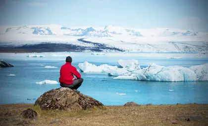 A visitor sitting by Jökulsárlón Glacier Lagoon in Iceland, admiring the floating icebergs with snow-capped mountains in the background.