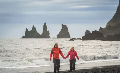 Smiling couple walks hand-in-hand on Reynisfjara black sand beach with Reynisdrangar basalt sea stacks in the background.