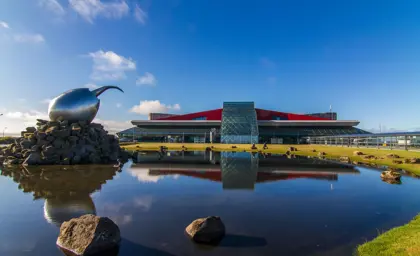 View of Keflavík International Airport in Iceland, featuring its modern architecture and iconic reflective sculpture by a calm pond on a sunny day.