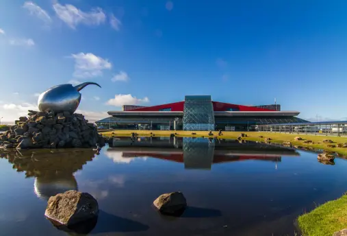 View of Keflavík International Airport in Iceland, featuring its modern architecture and iconic reflective sculpture by a calm pond on a sunny day.