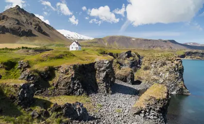 Arnarstapi's rugged coastline with dramatic cliffs, a quaint white house, and views of the Snaefellsnes Peninsula in Iceland, captured on a sunny day with blue skies.