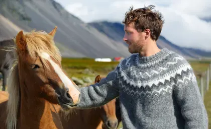 Man in traditional Icelandic lopapeysa sweater gently pets an Icelandic horse in a scenic mountain valley.