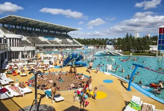 A vibrant scene at Laugardalslaug, Reykjavik’s largest swimming pool, featuring a spacious outdoor area with families enjoying the pools, waterslides, and sunlit seating under a bright summer sky.