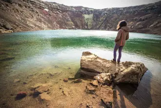 A woman standing on a rock overlooking the turquoise waters of Kerid Crater Lake in Iceland.