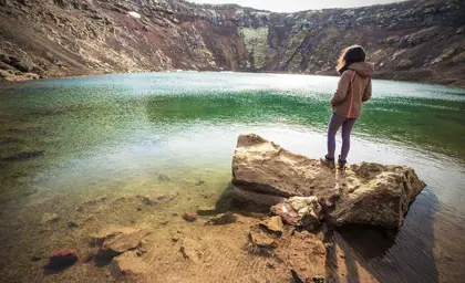 A woman standing on a rock overlooking the turquoise waters of Kerid Crater Lake in Iceland.