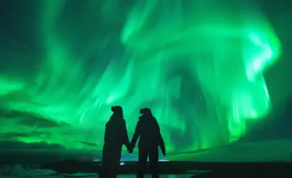 Couple holding hands under dazzling green northern lights in Iceland, reflecting in a still icy lagoon for a magical winter experience.