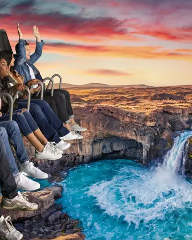 Group of visitors enjoying FlyOver Iceland's immersive experience, suspended over a scenic Icelandic waterfall landscape, with vibrant sunset skies in the background.