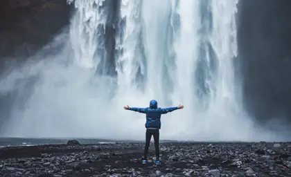 A person in a blue jacket stands with arms outstretched in awe before the powerful Skogafoss waterfall in Iceland, surrounded by mist and rocky terrain.