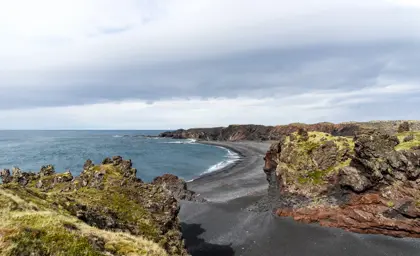 Panoramic view of Djúpalónssandur black sand beach in Iceland, featuring dramatic cliffs and the calm Atlantic Ocean under a cloudy sky, capturing the rugged beauty of Snæfellsnes Peninsula.