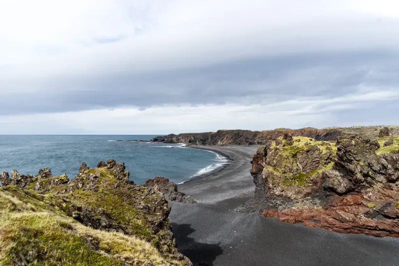 Panoramic view of Djúpalónssandur black sand beach in Iceland, featuring dramatic cliffs and the calm Atlantic Ocean under a cloudy sky, capturing the rugged beauty of Snæfellsnes Peninsula.