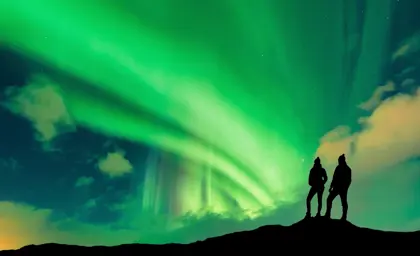 Silhouette of a young adventurous couple standing on a hill under a vivid green aurora borealis on a 4 day winter stopover tour in Iceland.