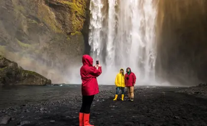 Friends in colorful rain jackets take a photo in front of Skogafoss waterfall with visible rainbow and mist in Iceland.