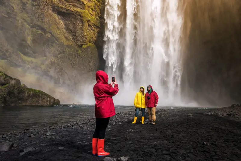 Friends in colorful rain jackets take a photo in front of Skogafoss waterfall with visible rainbow and mist in Iceland.