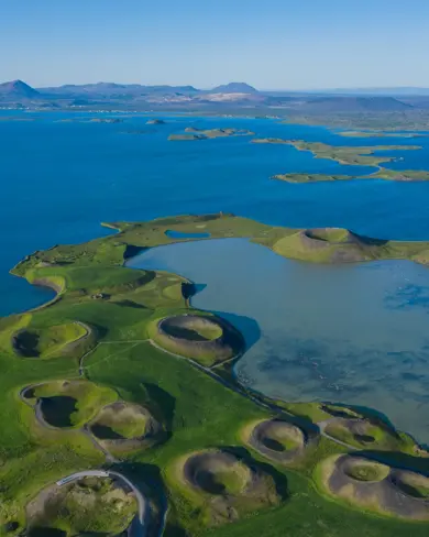 Aerial view of Skútustaðir pseudo craters on the shores of Lake Mývatn in North Iceland, showcasing unique volcanic landscape formations surrounded by lush green grass and pristine blue waters under a clear sky.