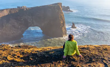 Young woman sitting on the cliff at Dyrhólaey, watching the ocean and sea arch during a winter sunset in South Iceland