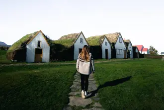Glaumbær turf house farm and museum behind a woman walking towards it surrounded by green grass and clear skies. 