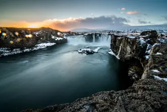 Goðafoss waterfall in winter in Iceland, with powerful turquoise water flowing through snow-dusted cliffs, capturing the dramatic winter landscape of this iconic site.