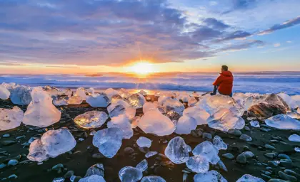 Person in red jacket sitting on ice chunk watching vibrant sunset at Diamond Beach with scattered ice formations on black sand