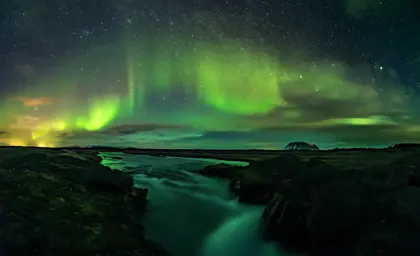 Vibrant Northern Lights reflecting over a flowing Icelandic river at night, a stunning view of Iceland's natural beauty during a scenic tour.