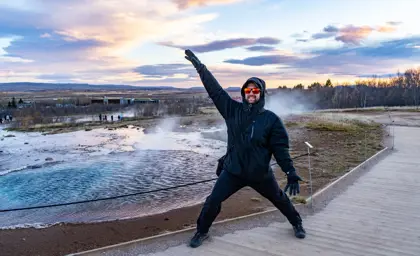 Joyful tourist in winter gear poses dramatically in front of Strokkur geyser erupting at the geothermal site in Iceland.