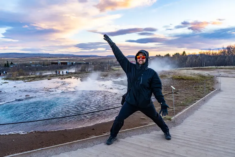 Joyful tourist in winter gear poses dramatically in front of Strokkur geyser erupting at the geothermal site in Iceland.