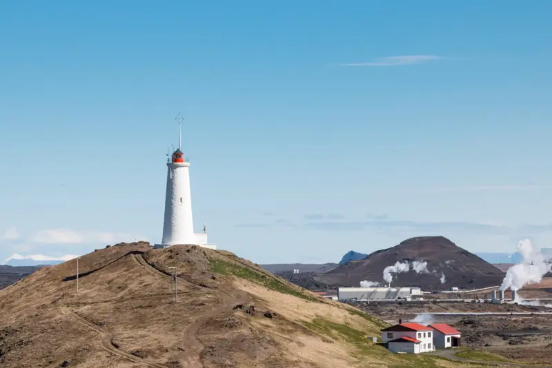 Reykjanesviti Lighthouse standing atop a grassy hill, surrounded by geothermal steam vents and rugged terrain, under a bright blue Icelandic sky.