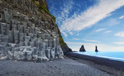 Reynisfjara Black Sand Beach with columnar basalt formations and Reynisdrangar sea stacks under a blue sky in Iceland, part of Travel Reykjavik tours.