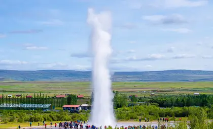 Geysir hot spring erupts in Iceland, surrounded by a crowd of onlookers against a backdrop of lush greenery and mountains. The iconic geyser displays a powerful burst of water under a bright blue sky, showcasing the geothermal activity that Iceland is famous for.