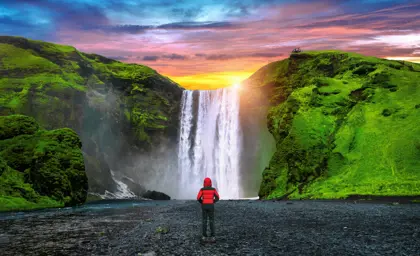 A person on a private south coast tour in a red jacket stands facing the majestic Skógafoss waterfall in Iceland during a vibrant sunset, with lush green cliffs on either side of the cascade.