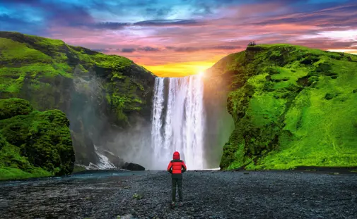 A person on a private south coast tour in a red jacket stands facing the majestic Skógafoss waterfall in Iceland during a vibrant sunset, with lush green cliffs on either side of the cascade.