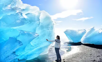 Tourist in Icelandic wool sweater touching glowing blue iceberg on Diamond Beach near Jökulsárlón glacier lagoon under bright winter sun.