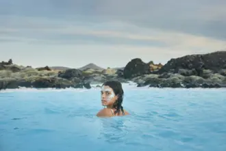 Woman with a white silica face mask relaxing in the Blue Lagoon, a geothermal spa in Iceland, surrounded by volcanic rocks and distant hills under a cloudy sky.