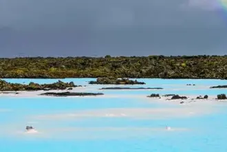 The blue waters of the Blue Lagoon surrounded by moss covered rocky black lava with a rainbow rising up above.