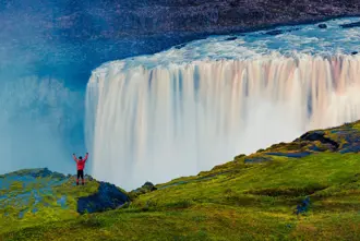 View of Dettifoss Waterfall where a hiker stands triumphantly in front of the most powerful waterfall in Europe, located in Iceland's Vatnajökull National Park, with mist rising and vibrant greenery surrounding the scene.