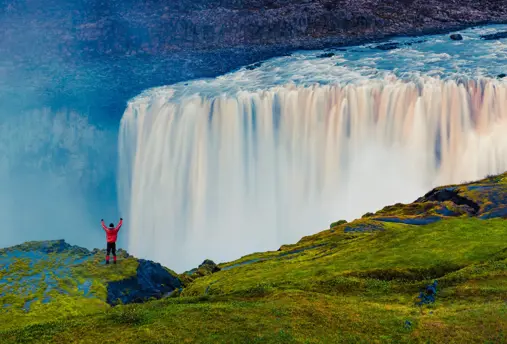 View of Dettifoss Waterfall where a hiker stands triumphantly in front of the most powerful waterfall in Europe, located in Iceland's Vatnajökull National Park, with mist rising and vibrant greenery surrounding the scene.