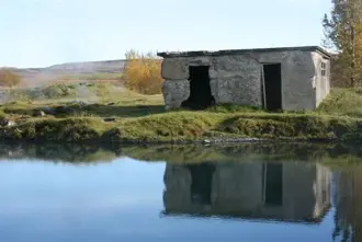 Historic stone bathhouse by the geothermal waters of the Secret Lagoon in Flúðir, Iceland, reflecting on the calm, blue pool surrounded by autumn foliage.