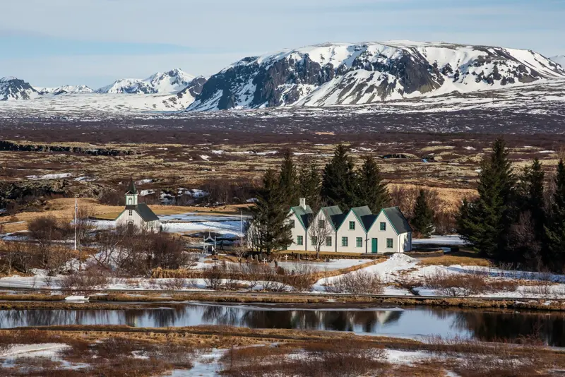 Scenic view of Þingvellir National Park with snow-covered mountains and traditional Icelandic church by the lake in early spring.